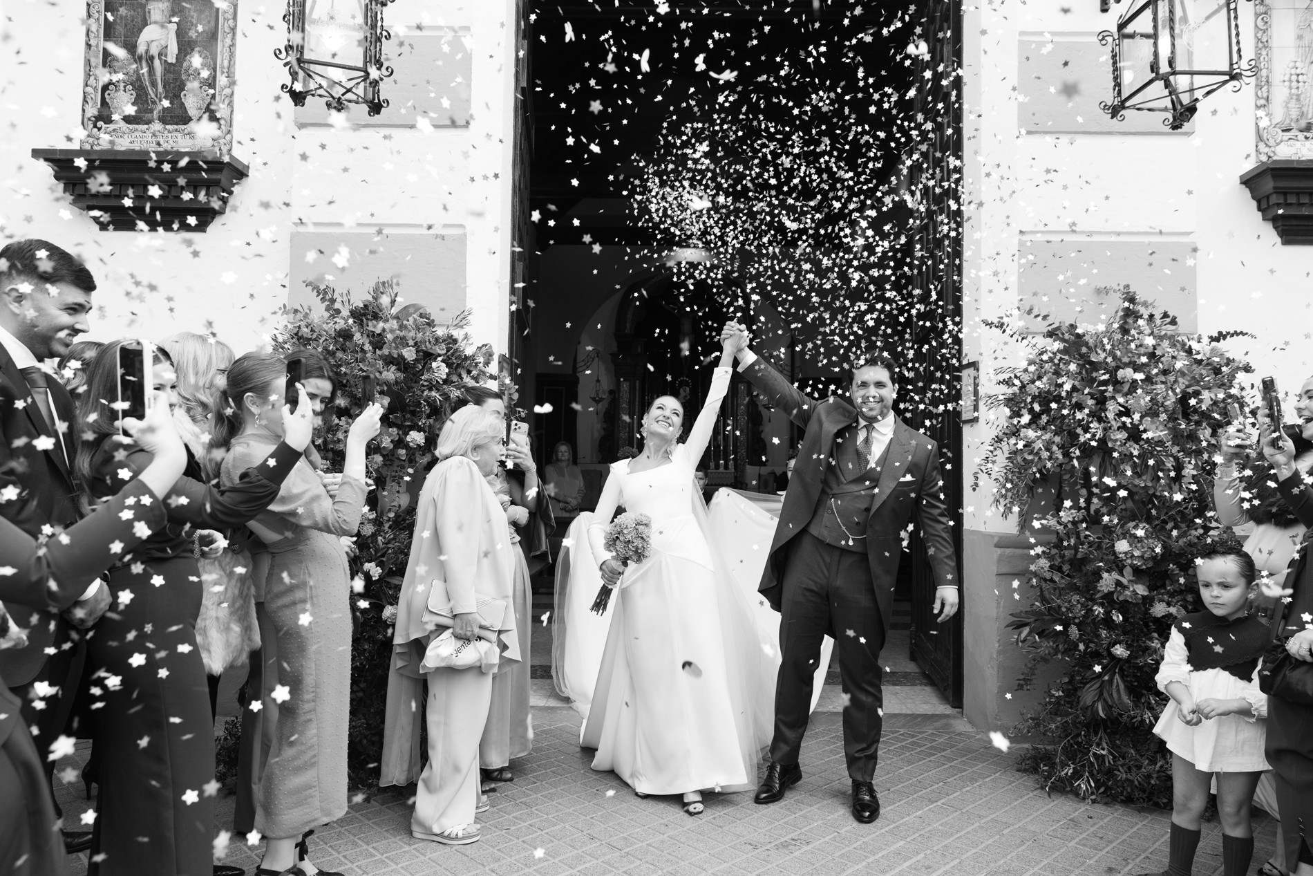 Los novios salen de la iglesia bajo una lluvia de confeti, ella con el brazo en alto, en blanco y negro